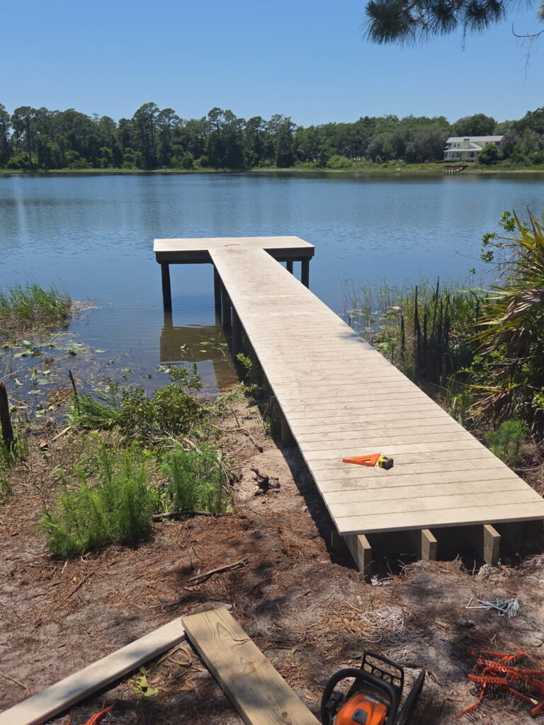 Unfinished wooden dock extends into a calm lake, with lumber and tools scattered on the muddy shore nearby.