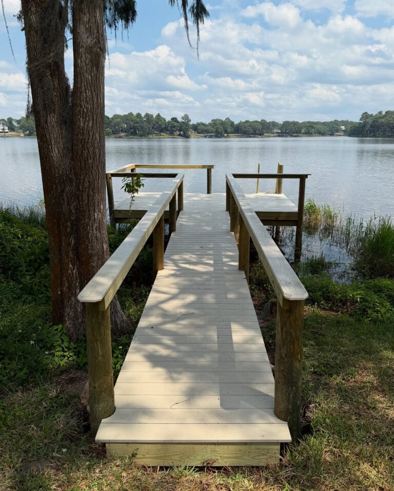 Wooden dock extending over a calm lake, with grassy banks and trees, under a partly cloudy sky.