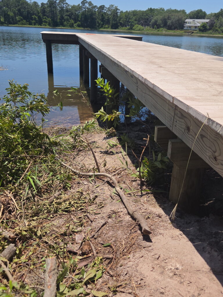 Wooden dock extending into a calm lake with a forested shore and a distant white house.