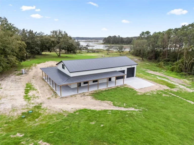 Aerial view of a modern white metal-roofed building with a long covered porch on a green, open field near a lake and surrounding trees.