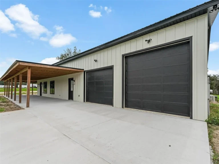 Industrial-style building with two large black roll-up garage doors under a wooden carport on a concrete lot