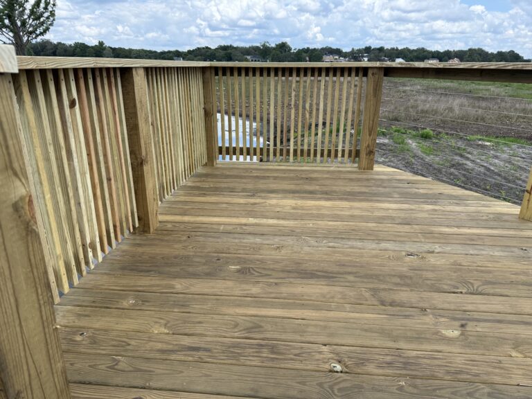 Wooden deck railing and floor overlooking a grassy field with distant houses under a cloudy sky.