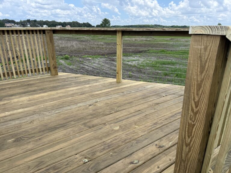 Wooden deck railing with metal cables overlooking a grassy, open field and distant houses under a cloudy sky.