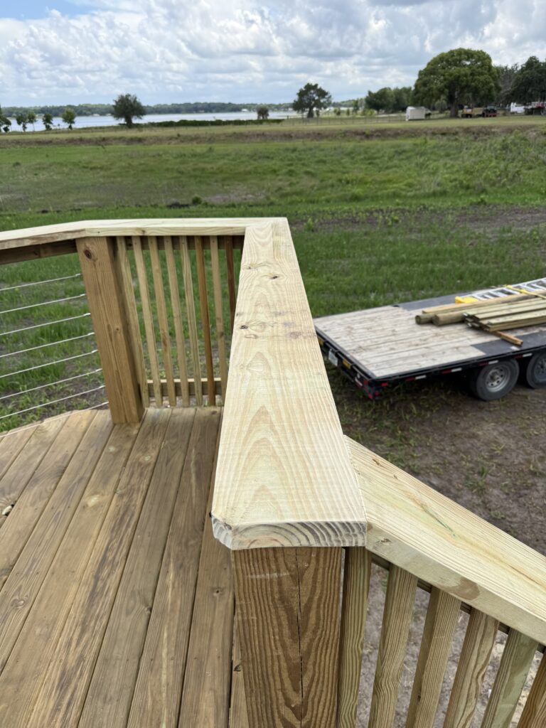 Close-up of a wooden deck railing and top rail with a trailer and building materials in a field beyond.