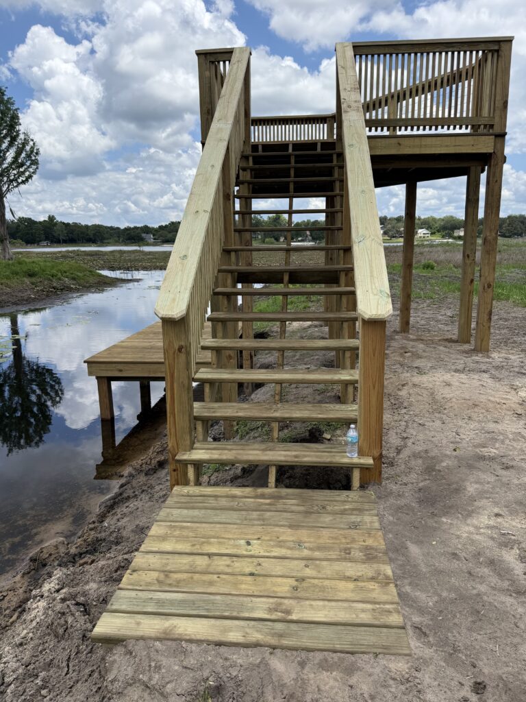 Wooden staircase and deck leading to a lakeside viewpoint, with water and blue sky in the background.