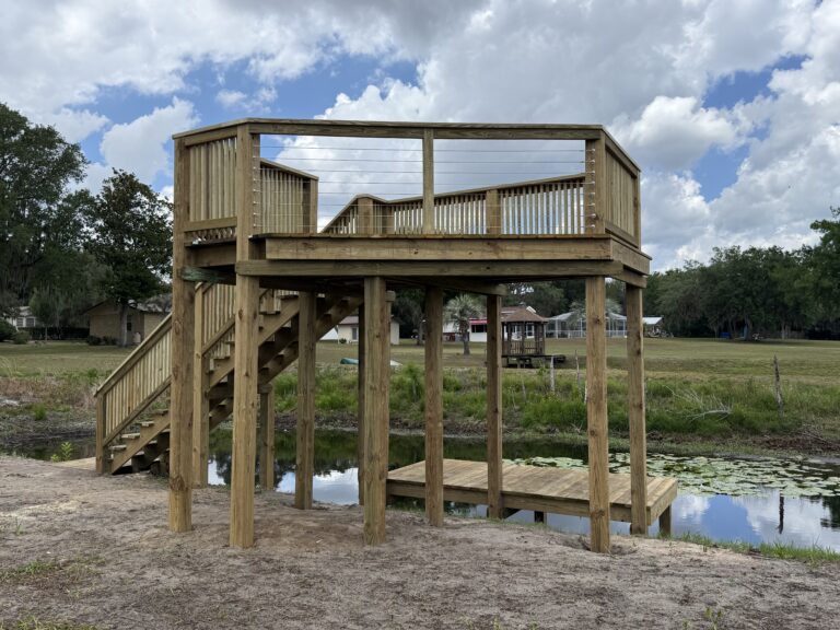 Two-level wooden deck on stilts above a small pond, with stairs on the left and cable railings, in a park with trees and houses in the background under a cloudy sky.