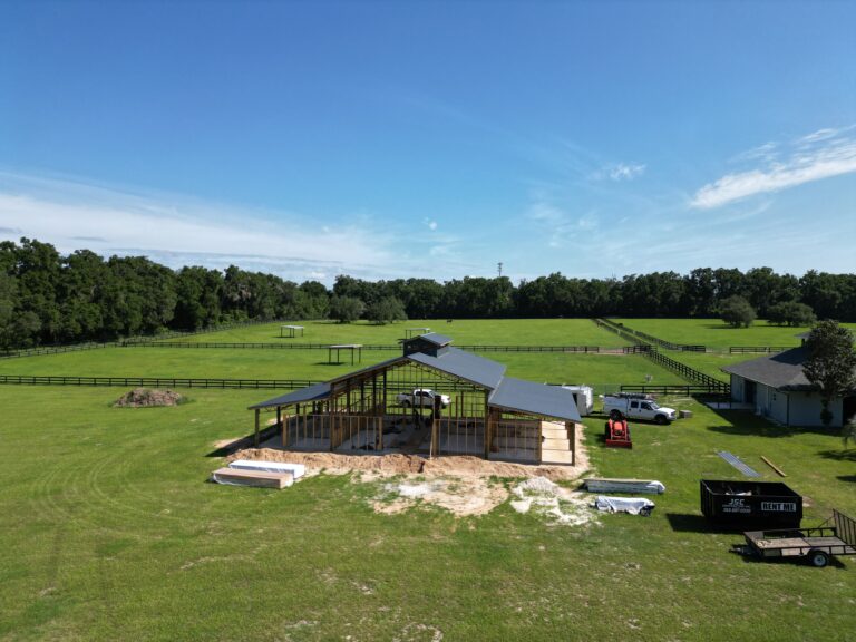 Open field with a partially built wooden barn under construction, construction materials and equipment nearby, under a clear blue sky