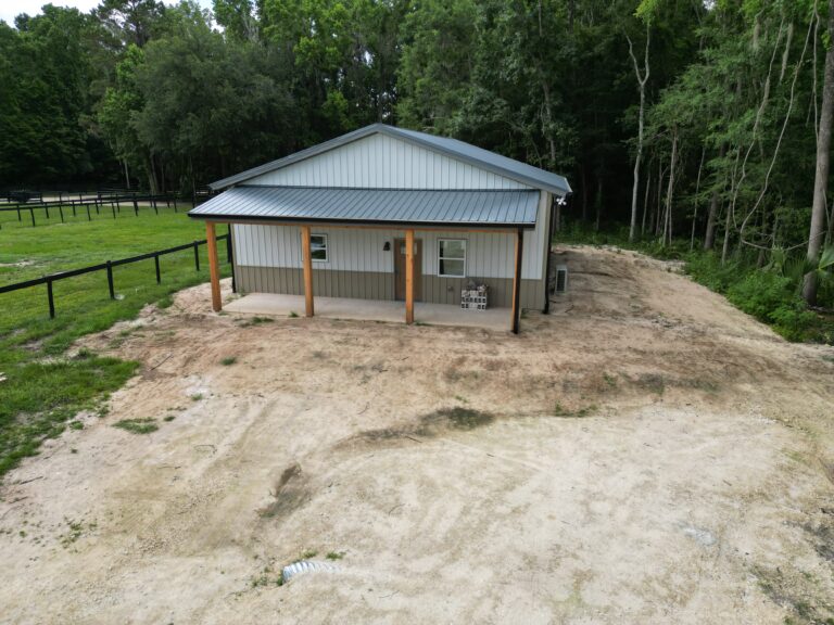 Small metal-roofed building on a dirt lot, with a wooden porch posts and a surrounding wooden fence, backed by trees and woods.