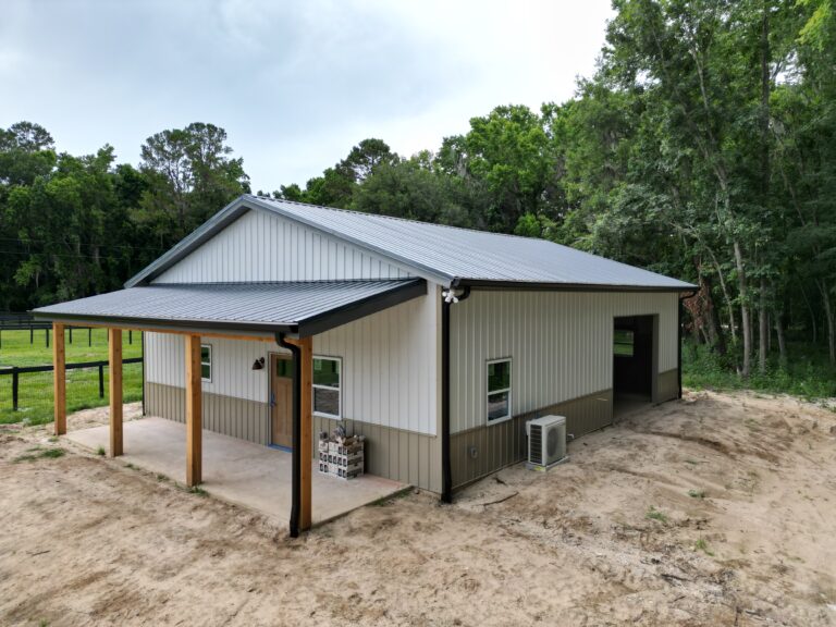Metal-clad building with a gray corrugated roof, wooden porch posts, and dirt yard with trees in the background.